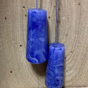 May include: Two blue, cylindrical pull chains with a marbled effect hang against a wooden background. The chains are attached to the top of the cylinders with a silver string. Several seashells are scattered at the bottom.