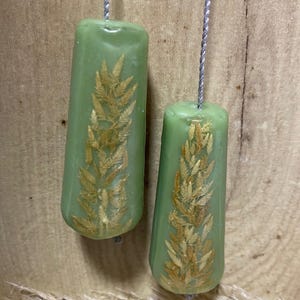 May include: Two light green, cylindrical candle holders with dried fern designs encased within. Each holder is suspended by a silver-grey string. The background is a light brown wooden surface.
