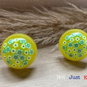 May include: Two yellow round knobs with a floral design. The knobs have green and white flowers on a yellow background. The knobs are on a light brown surface.