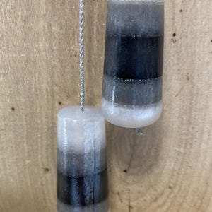 May include: Two hanging, cylindrical candles with a gradient of colours. The top is white, transitioning to grey, then black, and back to grey. The candles are suspended by a silver string. The background is a light brown wooden surface.