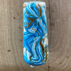 May include: A cylindrical pull with a swirling pattern of blue, white, and brown. It is suspended by a silver cord. The background is a wooden surface.