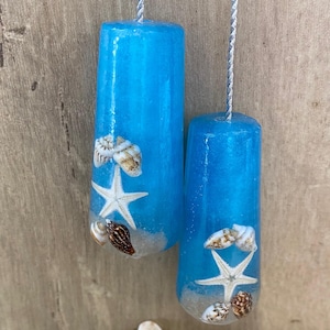 May include: Two cylindrical blue resin ornaments hanging from silver cords, each embedded with white starfish and small seashells at the base. The resin has a translucent, shimmering quality, and a collection of loose seashells rests below them.