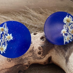 May include: Two round blue resin earrings with white pressed flowers. The earrings are approximately 2.5 cm in diameter.