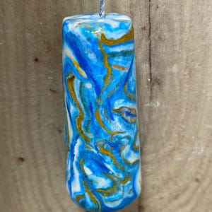 May include: A cylindrical pendant featuring a marbled pattern of bright blue, white, and metallic gold swirls. The object has a smooth, glossy finish and is suspended from a thin, light grey twisted cord that passes through the centre.