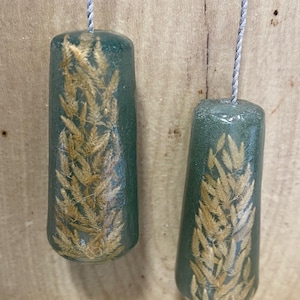 May include: Two teal light pulls, each with a wheat stalk design encased within. They are suspended by silver cords. The background is a light brown wooden surface. The pulls are approximately 5 cm in length.