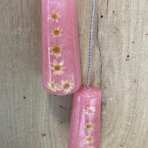 May include: Two pink resin light pull chains with embedded dried flowers. The pulls are cylindrical with a slight taper and are suspended by a silver cord. The background is a light brown wooden surface.
