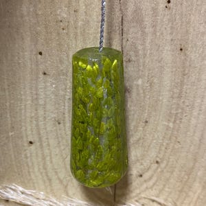 May include: A green, cylindrical light pull with a silver cord. The pull is made of a translucent material with a pattern of green leaf-like inclusions. The pull is suspended against a wooden background.