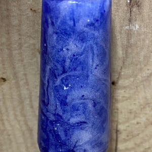 May include: A cylindrical pendant with a smooth, glossy finish featuring a marbled pattern of deep royal blue and shimmering white swirls. The pendant is suspended from a thin, silver-coloured cord that passes through a hole at the top centre.