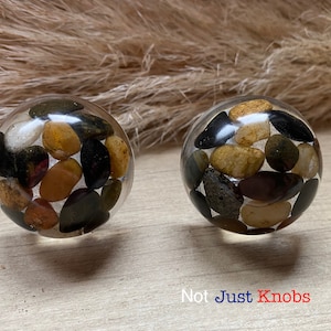 May include: Two round clear resin knobs with embedded small stones in shades of black, brown, and beige. The text "Not Just Knobs" is visible below the knobs.