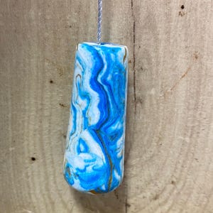 May include: A cylindrical light pull with a marbled design. The pull features a swirling pattern of blue, white, and gold tones. A silver cord is attached to the top of the pull, which hangs against a wooden background.