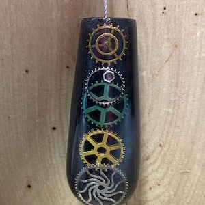 May include: Black, teardrop-shaped pull with embedded gold, silver, and green gears. A silver cord is attached at the top. The gears are arranged vertically on the front of the pull. The background is a light brown wooden surface.