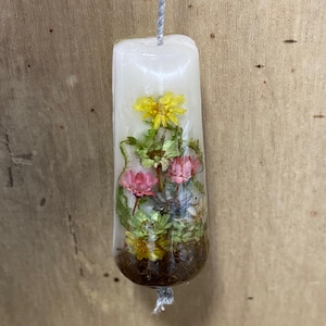 May include: A cylindrical white wax ornament featuring embedded dried flowers in shades of yellow, pink, and green, with a dark brown base. A thin silver string runs vertically through the centre of the wax, extending from the top and bottom.