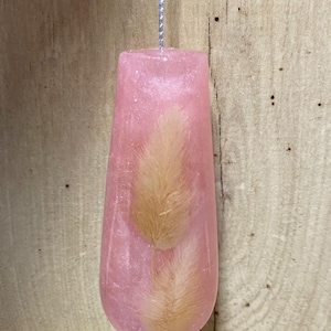 May include: A pink resin pull with a silver string. The pull is teardrop-shaped and contains two dried, fluffy, tan-coloured bunny tail grass stems. The background is a light brown wooden surface.
