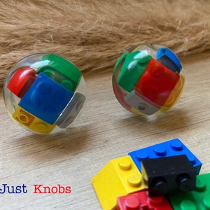 May include: Two clear plastic spheres filled with colourful Lego blocks. The spheres are sitting on a wooden surface. The text "Not Just Knobs" is visible below the spheres.