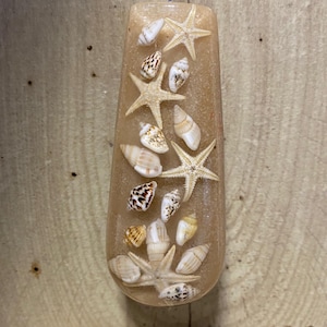 May include: A teardrop-shaped resin pendant in a light brown hue, featuring embedded seashells and starfish. It hangs from a thin, twisted cord. The background is a wooden surface, with loose seashells scattered below.