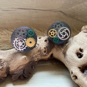 May include: Two black resin rings with silver and gold gears and green accents. The rings are on a piece of driftwood.