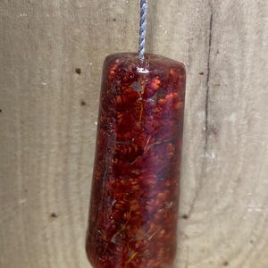 May include: A cylindrical, reddish-brown resin object with embedded floral elements, suspended by a grey string. The resin is translucent, revealing the intricate details of the dried flowers within. Likely a decorative pull or pendant.