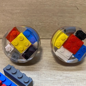 May include: Two clear, round resin objects each containing colourful toy building blocks. The blocks are red, yellow, blue, black, and white. Additional loose blocks are visible at the bottom of the image.
