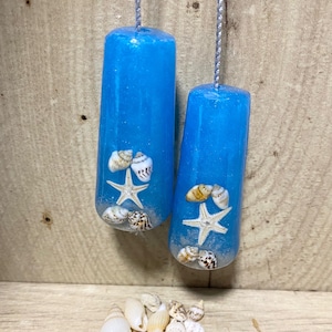 May include: Two blue, cylindrical pull chains with embedded seashells and starfish, suspended by silver cords. A collection of seashells rests below on a wooden surface. Coastal theme.