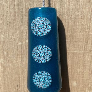 May include: A cylindrical teal blue pendant with a subtle glitter finish, featuring three circular inlays decorated with a geometric floral pattern in shades of blue, white, and red. The pendant is suspended from a thin, silver-coloured cord.