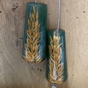 May include: Two tapered, dark green resin pendants, each containing a preserved golden wheat stalk. The pendants are suspended by thin, silver-coloured braided cords. The resin has a smooth, glossy finish, and the wheat stalks are centred within.