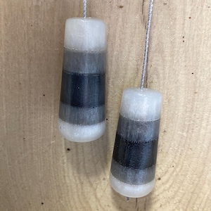 May include: Two cylindrical light pull cords with a gradient design. The top and bottom sections are white, with a dark grey and black band in the middle. The cords are silver and hang against a wooden background.