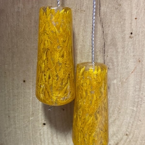May include: Two cylindrical, yellow resin light pulls with a textured, fern-like design. Each pull is suspended by a silver-coloured cord. The background is a light brown wooden surface.