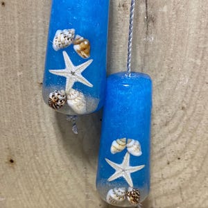 May include: Two cylindrical blue resin light pulls featuring embedded white starfish and various small seashells, suspended by silver braided cords. A collection of loose, assorted seashells is arranged at the base of the two hanging objects.