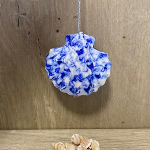 May include: A blue and white marbled seashell ornament suspended by a silver string. A collection of small seashells rests on a wooden surface below. The ornament is a decorative item with a coastal theme.