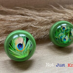 May include: Two green round cabinet knobs featuring a peacock feather design. The knobs are displayed on a wooden surface with a tan grass-like plant in the background. The text "Not Just Knobs" is visible at the bottom of the image.