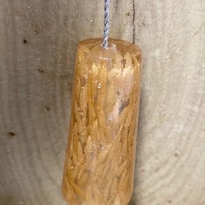 May include: A tapered, cylindrical pendant made of clear resin containing embedded golden-brown dried wheat stalks. The object is suspended from a thin, twisted grey cord, showcasing a smooth, glossy finish and a natural, organic texture throughout.