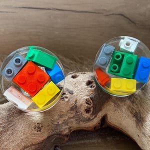 May include: Two round resin coasters with embedded colourful Lego bricks. The coasters are clear and show the Lego bricks in various colours, including red, blue, green, yellow, and grey.