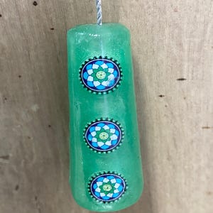 May include: A cylindrical green pendant suspended by a silver-coloured cord. The pendant features three circular inlays arranged vertically, each displaying a blue, white, and green floral pattern surrounded by a small beaded metal border.