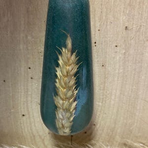 May include: A teal-coloured, teardrop-shaped pull with a dried wheat stalk encased within. The pull is suspended by a silver cord. The background is a light brown wooden surface.