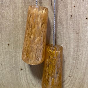 May include: Two amber-coloured resin light pull cords with a feathery, golden interior design. The pulls are suspended by silver cords against a wooden background, creating a warm, natural aesthetic.
