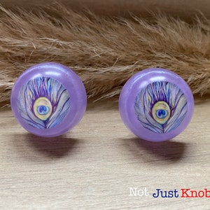 May include: Two purple round cabinet knobs with a peacock feather design. The knobs have a shiny, iridescent finish.