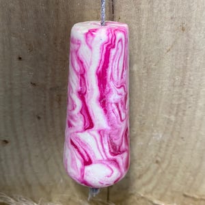 May include: A cylindrical pull with a pink and white marbled pattern, hanging from a grey string. The pull is set against a wooden background.