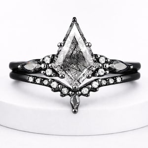 May include: A black engagement ring set with a large, kite-shaped, clear gemstone with black inclusions. The set includes a matching band with small, clear gemstones and black metal accents. The rings are displayed on a white surface.