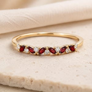 May include: A gold band ring featuring an alternating pattern of marquise-cut deep red stones and round brilliant-cut clear stones. The stones are set along the front arc of the thin, polished gold metal band, creating a delicate and symmetrical design.
