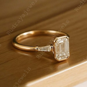 May include: A gold ring with a large emerald-cut diamond as the centerpiece. Two smaller baguette diamonds flank the main stone. The ring is set on a simple, polished gold band, showcasing a classic and elegant design.