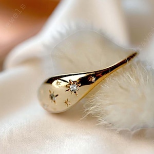 May include: A gold-toned ring featuring a smooth, teardrop-shaped surface adorned with multiple engraved star motifs. Several of these star designs are accented with small, clear, sparkling stones embedded at their centres, creating a celestial pattern.