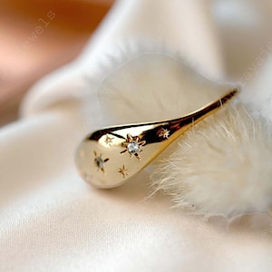 Puede incluir: Un anillo dorado con forma de lágrima, adornado con pequeños diseños en forma de estrella y una gema central brillante. El anillo se presenta sobre un fondo suave y blanquecino.