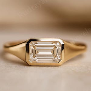 May include: A gold ring with a rectangular, emerald-cut diamond. The diamond is set in a bezel setting, and the band is smooth and polished. The ring is displayed on a neutral background.