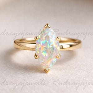 May include: A gold ring featuring a marquise-cut, iridescent gemstone. The gemstone is set with four prongs and has a pointed top and bottom. The band is a simple, polished gold. The ring is set against a soft, neutral background.