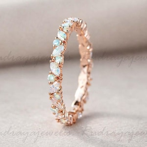 Pode incluir: Anel de ouro rosa com uma faixa contínua de pedras preciosas de opala e claras alternadas. As pedras de opala são branco leitoso e azul claro. O anel está sobre um fundo neutro.