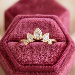 Könnte beinhalten: Ein goldener Ring mit Kronen-Design, mit birnenförmigen und runden Diamanten. Der Ring befindet sich in einer Samt-Schmuckschatulle. Die Diamanten funkeln auf dem Goldband und erzeugen einen luxuriösen Look.