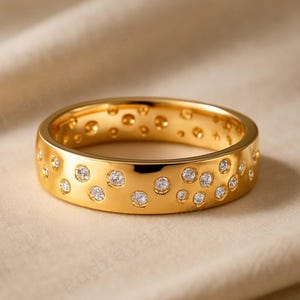 May include: A gold-colored ring with a band featuring a pattern of small, evenly spaced, clear gemstones. The ring has a polished finish and is set against a neutral background.