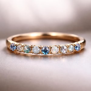 Op de afbeelding: Een gouden bandring met een rij kleine, kleurrijke edelstenen. De edelstenen zijn in tinten blauw, wit en groen. De ring is geplaatst tegen een zachte, neutrale achtergrond, die het delicate ontwerp benadrukt.