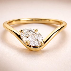 Op de afbeelding: Een gouden ring met een V-vormige band en een grote, ovaal geslepen diamant als middensteen. De diamant wordt op zijn plaats gehouden door vier pootjes. De ring staat tegen een zachte, crèmekleurige achtergrond.