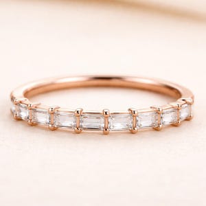 May include: Rose gold ring with a band set with a row of rectangular, clear gemstones. The ring's band has a warm, metallic hue, and the gemstones are closely set. The ring is shown against a soft, neutral background, emphasizing its elegant design.